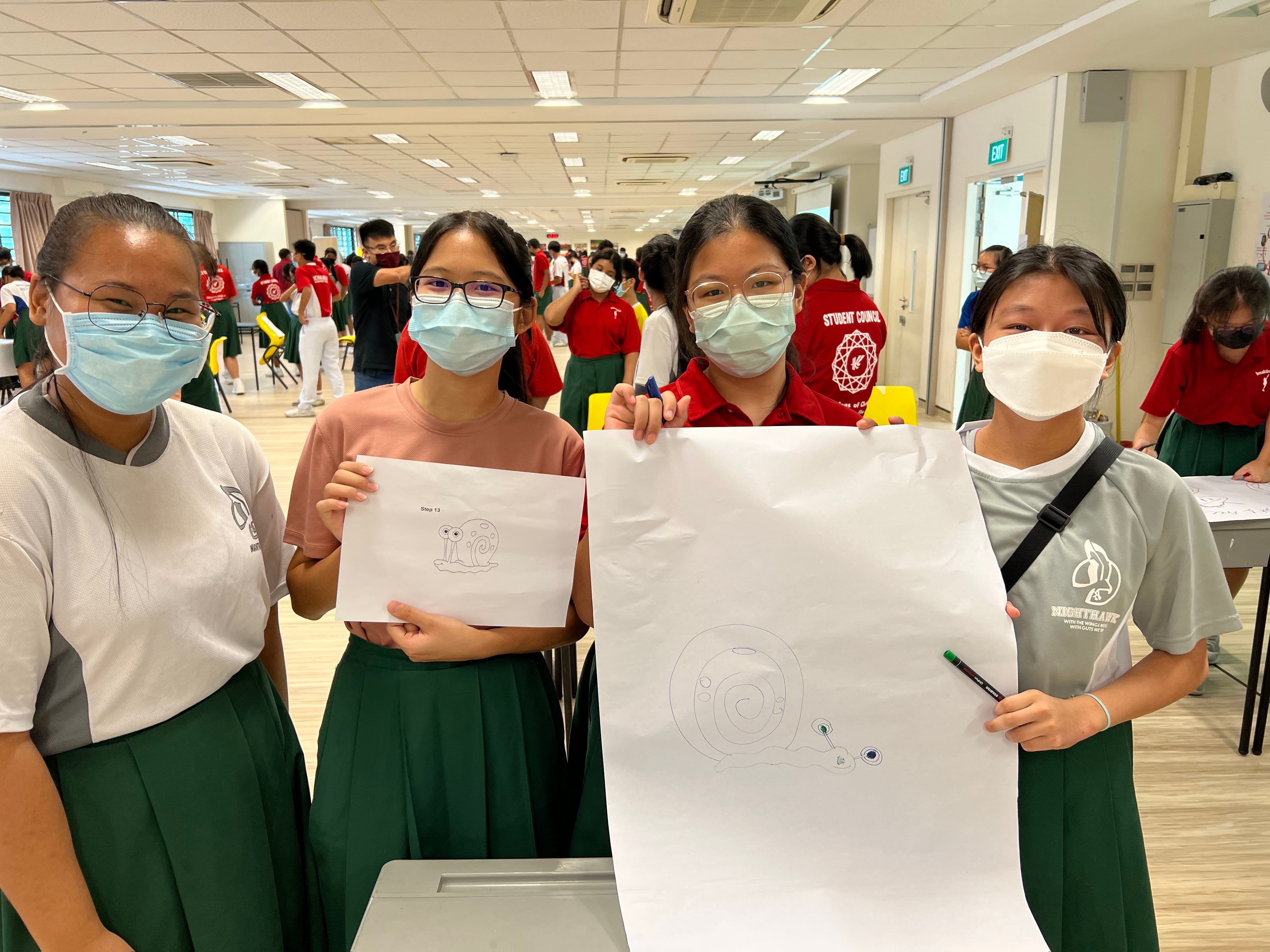 Four masked students in school uniforms hold up papers with snail drawings in a busy indoor hall.